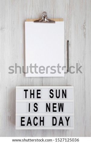 'The sun is new each day' words on a modern board, clipboard with blank sheet of paper on a white wooden surface, view from above. Top view, overhead, flat lay. 