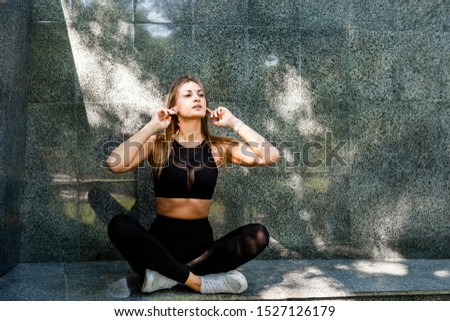 Young sporty woman in black sportswear posing over marble wall background. Sporty girl with wireless headphones relaxing and listening to music or podcast.