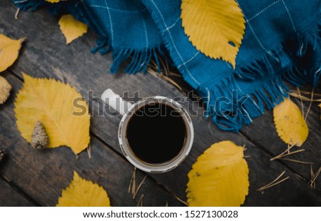 On a old wooden table in the autumn park is a cup of tea-coffee. A blue warm plaid scarf is scattered with yellow leaves and pine cones. Top view, blurred. Autumn warm dark mood, copy space