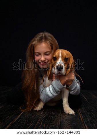 A very cute and beautiful little girl of ten years of age indulges in a small beagle dog sitting on a dark wooden plank floor. The dog tries to knock the child to the floor, bites the girl .
