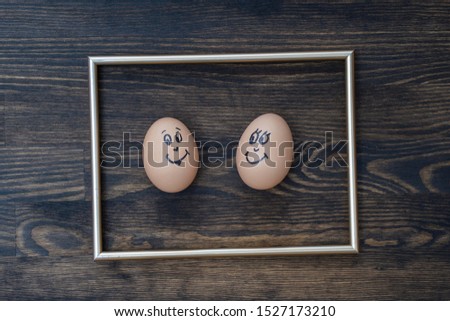 Picture golden frame and two funny eggs smiling on dark wooden wall background, close up. Eggs family emotion face portrait. Couple eggs with happy face for love concept