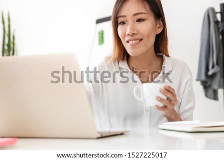 Attractive smiling young asian businesswoman working while sitting at the desk in office, holding cup of coffee, while using laptop computer