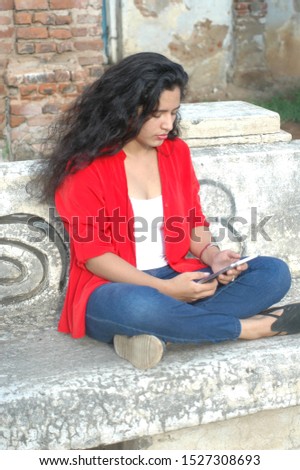 Young wavy hair sitting on a semento bench with its feet between loops, looking straight at her tablet while smiling, wearing a red shirt over a white shirt and blue trousers. Photo portrait.