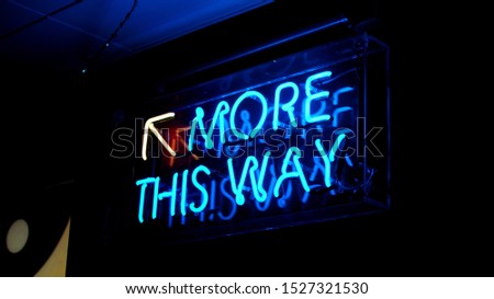 A bright blue fluorescent 'More This Way' neon sign with a yellow arrow pointing upwards. The neon sign illuminates its surroundings making this a key feature to the growing establishment
