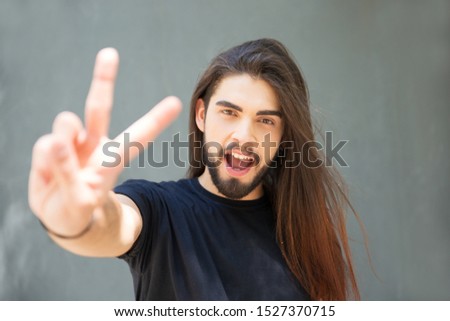Excited long haired guy showing victory gesture. Handsome bearded young man standing over grey background, making peace sign with hand and fingers. Gestures concept