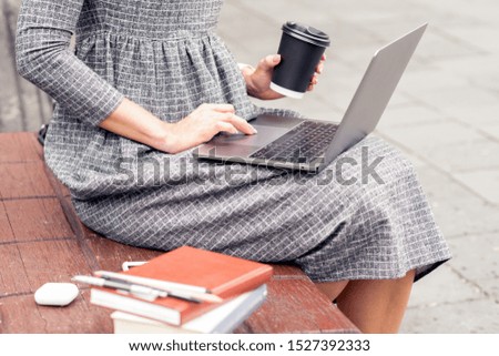 Close up of young woman's hand holding a cup of coffee while using a laptop on the bench. Close up of a female student works on a laptop outdoors.