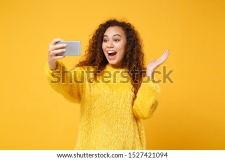 Excited young african american girl in fur sweater posing isolated on yellow orange wall background. People lifestyle concept. Mock up copy space. Doing selfie shot on mobile phone, spreading hands