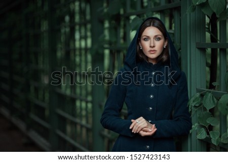 Poetic beautiful brown hair brunette woman or girl in old vintage classic dress coat looks camera by green eyes and holds hands together laying on backyard garden park iron forged fence Royalty-Free Stock Photo #1527423143