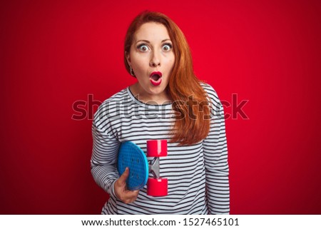 Young redhead student woman holding skate over red isolated background scared in shock with a surprise face, afraid and excited with fear expression