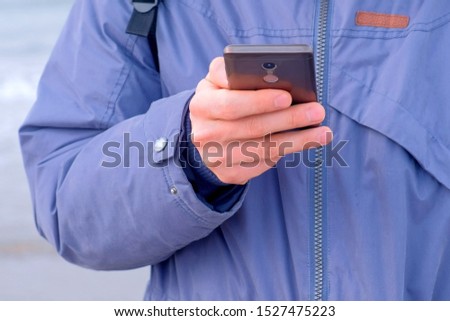 Man typing message on smartphone in coastal town at sea in winter, hands clseup. Man traveller freelancer blogger is writing post on phone. Tourist travel journey trip with backpack.