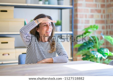 Middle age senior woman sitting at the table at home very happy and smiling looking far away with hand over head. Searching concept.