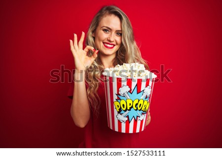 Young beautiful woman wearing t-shirt eating popcorn over red isolated background doing ok sign with fingers, excellent symbol