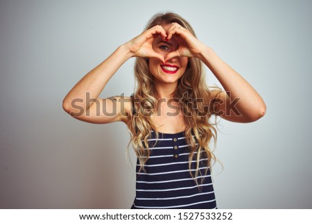 Young beautiful woman wearing stripes t-shirt standing over white isolated background Doing heart shape with hand and fingers smiling looking through sign