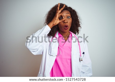 African american doctor woman wearing  pink stethoscope over isolated white background doing ok gesture shocked with surprised face, eye looking through fingers. Unbelieving expression.