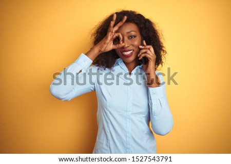 Young african american woman talking on the smartphone over isolated yellow background with happy face smiling doing ok sign with hand on eye looking through fingers