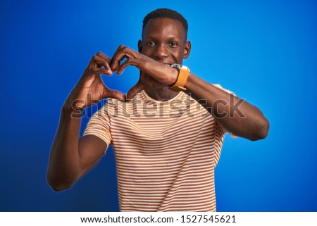 African american man wearing striped casual t-shirt standing over isolated blue background smiling in love showing heart symbol and shape with hands. Romantic concept.