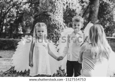 Young blonde woman walks with children - a little boy and a girl - in the park and photographs them on her cell phone. Children dressed as white-winged angels