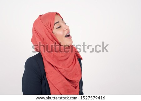 Joyful Muslim woman having fun and laughs at good joke or sings wears casual clothes, standing against gray wall. Happy woman with long hair poses inddoors.
