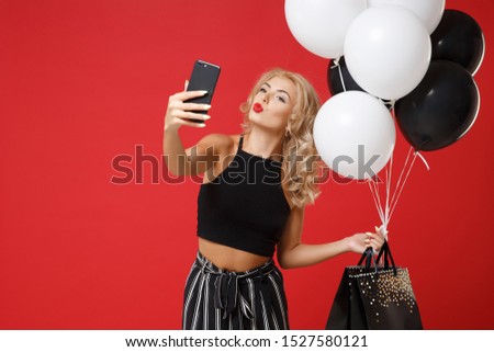 Pretty woman in black clothes posing isolated on red background. Shopping discount sale concept. Mock up copy space. Hold air balloons, package bag with purchases, doing selfie shot on mobile phone