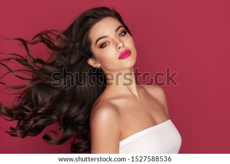 Beautiful Brunette Woman Portrait with brown eyes and Healthy Long Shiny Wavy black hair. Volume shampoo. Attractive Beauty model with professional make up against red background Royalty-Free Stock Photo #1527588536