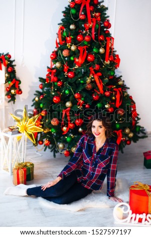 The girl in stylish clothes sitting on the floor near the christmas tree and looking at the camera lens