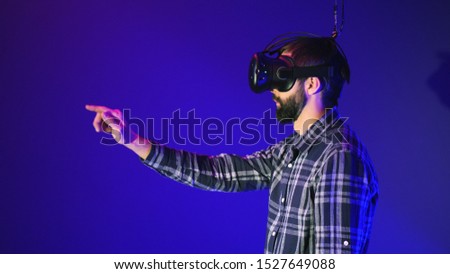 Bearded man using virtual reality headset at modern office with blue background, turns over, clicks, man work in VR, gesticulating by hands, side view