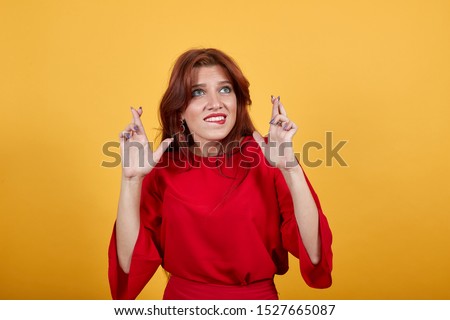 Charming Europen lady crossing her fingers on both hands and having unsured expression of face. Beautiful girl in red dress standing on yellow background having magnetic gray eyes.
