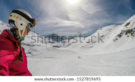 A snowboarder taking a selfie while going down the slope in Mölltaler Gletscher, Austria. High mountains surrounding the girl wearing colourful snowboard outfit. Girl wears helm for the protection