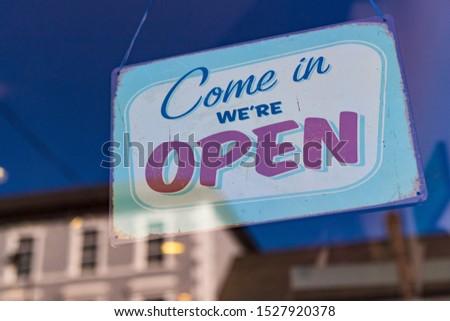 Come in we're open sign in a store window, blue sky and building reflected in the glass