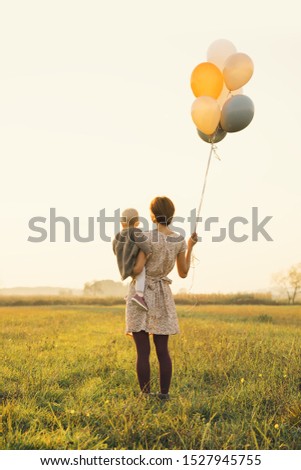 Young mother and baby girl with balloons in sunlight at sunset on nature outdoors. Mother and child in meadow in early autumn. Photo of leisure, dreams, holidays, family values, sustainable lifestyle.