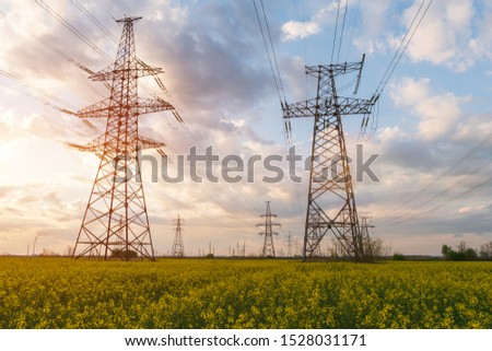 Power lines and high-voltage lines against the backdrop of blooming oilseed rape on a summer day. Green energy. Transmission of electricity by means of supports through agricultural areas.