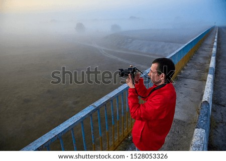 A man on a bridge over the river that photographs a sunrise on a foggy early morning.