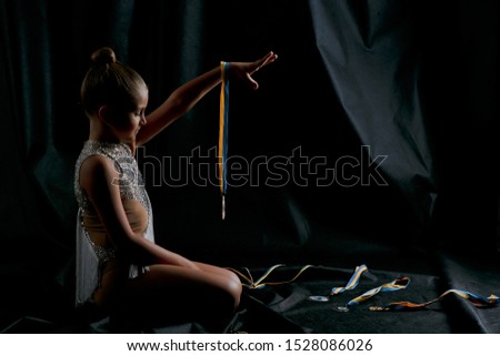 Beautiful girl teenager gymnast in a white sport dress looking at her medals on black background, medal hanging on her hand, side view