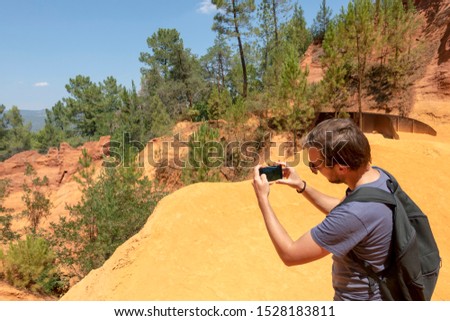 Young handsome man in mountains, caucasian male traveler take a picture of the Ochre Trail in Roussillon, Hiking path in orange ocher cliffs and green forest in Provence, Southern France