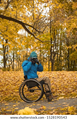 disabled person with paralyzed legs after spinal cord injury in a wheelchair takes pictures of nature in autumn park, daylight