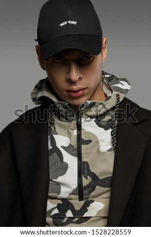 Close-up portrait shot of a short-haired European man dressed in a dazzle hooded top, a black jacket and a black baseball cap with an embroidered black lettering "New York".