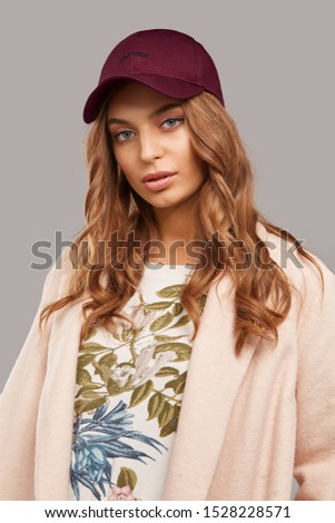 Medium close-up shot of blonde European lady with wavy hair in a white top with floral print, a pale pink jacket and a burgundy baseball cap with an embroidered black lettering "New York".