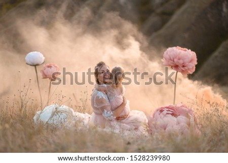 A little girl hugs mom sitting in a field surrounded by unreal big pink decorative flowers.