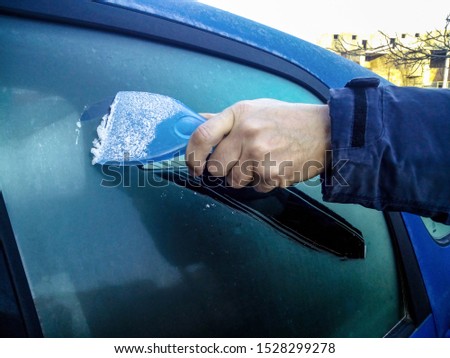 Man scrapes hoarfrost with a plastic scraper from the side window of a blue car. Hand without gloves in the cold. High-rise building in the distance. Close-up. Selective focus. Copy space. Royalty-Free Stock Photo #1528299278