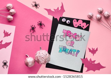 Creative paper craft Halloween flat lay in pink, light purple and black. Top view with stack of cards, bats, chocolate eyes and pink decorative pumpkins. Text "Boo" and "Trick or treat".