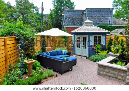 Contemporary with traditional elements, this beautiful small urban backyard garden features a seat wall, red brick paver herringbone pattern patio, natural stone steps and relaxing furniture. Royalty-Free Stock Photo #1528368830