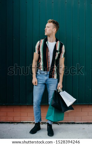A handsome young blond man in a striped shirt and light jeans is posing with bags near the green wall on Black Friday