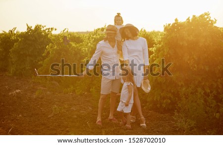 family, leisure and technology concept - happy mother, father and daughters having picnic and taking picture by smartphone on selfie stick at summer park. Sunset background 