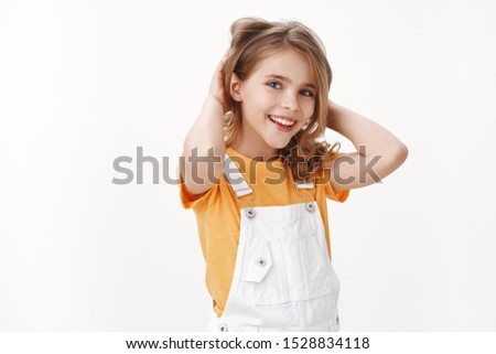 Cute pretty happy blond girl feeling upbeat, enjoy summer holidays, free from homework, stand yellow t-shirt and overalls, touch hair, smiling pleased, finally get rid of head lice, white background