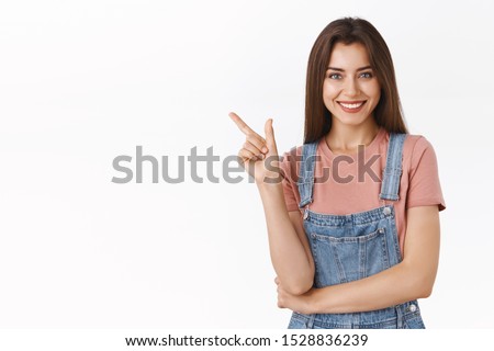 Cheerful, confident stylish modern woman in dungarees, t-shirt pointing upper left corner, laughing and smiling carefree as give hint where buy clothes during black friday, white background Royalty-Free Stock Photo #1528836239