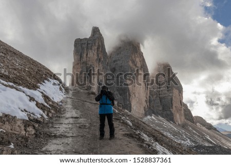 A woman in a blue jacket, a tourist, a photographer on a trek around Tre Cime Lavaredo in the Italian Dolomites, with mountains and a blue sky with clouds in the background.