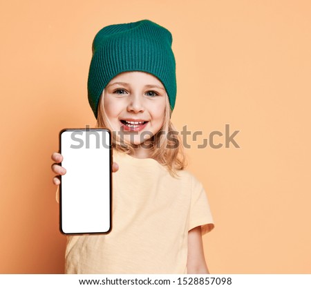 Smiling little girl kid in green modern winter hat showing white blank screen for text space of new popular mobile phone on light yellow background