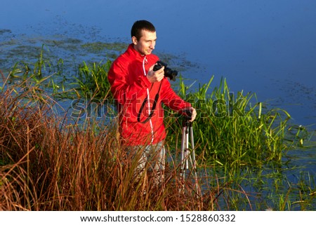 A man stands in a thicket of grass and photographs the river in the early autumn morning.