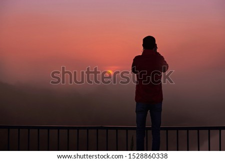 A silhouette of a man on a bridge over the river that photographs a sunrise on a foggy early morning.