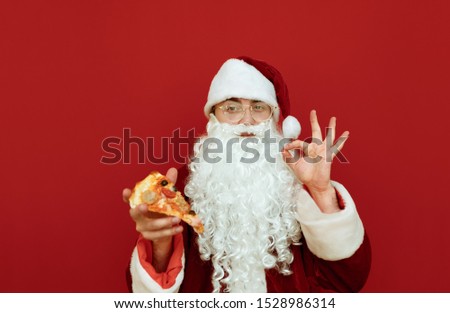Handsome Santa with a slice of pizza in her hand stands on a red background, looks in camera and shows her thumbs sign OK. Man in a Santa suit eats a pizza. Pizza for Christmas